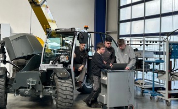 © VDMA: Generalprobe Bauma Das Bild zeigt einen gelben Teleskoplader mit geöffnetem Motorraum in einer hellen Werkstatt, umgeben von drei Personen, die auf einen Laptop schauen.