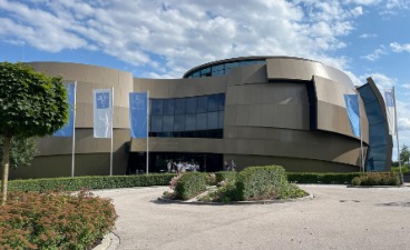 ESO Supernova Ein großes graues Gebäude in futuristischem Stil mit wehenden Fahnen davor und Bäumen links und rechts