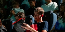 Ein Schüler sitzt im Planetarium und schaut Lächelnd auf sein Geschenk in der Hand, während um ihn herum weitere Besucher gespannt warten.