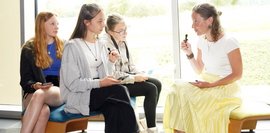 Drei Schülerinnen sitzen einer Frau gegenüber, die Frau spricht in ein Mikrofon und die Mädchen hören ihr zu.