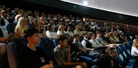 Ein Planetarium ist mit Menschen gefüllt, die in Reihen sitzen und gespannt nach vorne schauen.