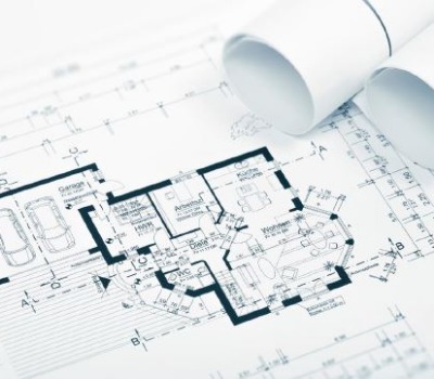 Das Bild zeigt einen detaillierten Gebäudegrundriss, auf dem Raumaufteilungen und Maße zu sehen sind, daneben liegen mehrere zusammengerollte Baupläne.
