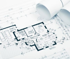 Auf dem Bild ist ein detaillierter architektonischer Grundriss zu sehen, der verschiedene Räume, Maße und bautechnische Angaben zeigt; daneben liegen mehrere zusammengerollte Baupläne, die den Eindruck einer professionellen Planungs- oder Entwurfsphase vermitteln.
