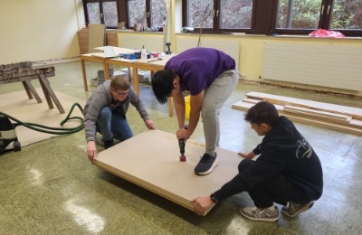 Das Bild zeigt drei Jugendliche in einer Werkstatt, die gemeinsam eine große Holzplatte bearbeiten, wobei einer mit einer Bohrmaschine arbeitet und die anderen die Platte stabilisieren.