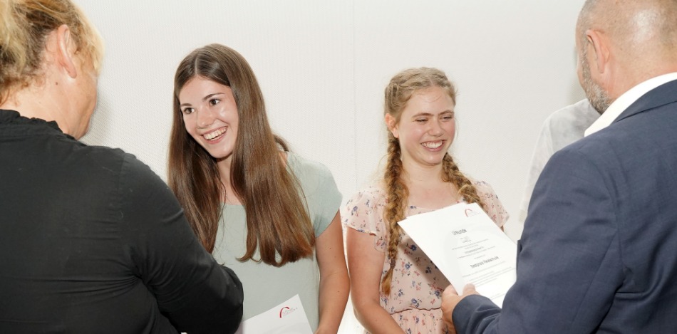 Abschlussveranstaltung 2023 Zwei lächelnde Mädchen, die von einer Frau und einem Mann etwas überreicht bekommen, die der Kamera den Rücken zugedreht haben