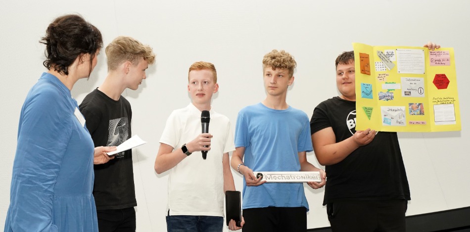 Abschlussveranstaltung 2023 Vier Schüler stehen mit einer Frau da, ein Junge hält ein Mirko und ein andere Junge hält ein Plakat nach oben