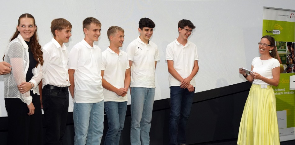 Eine Gruppe von sechs Jugendlichen steht in einer Reihe, eine Frau steht ihnen gegenüber und spricht zu ihnen.