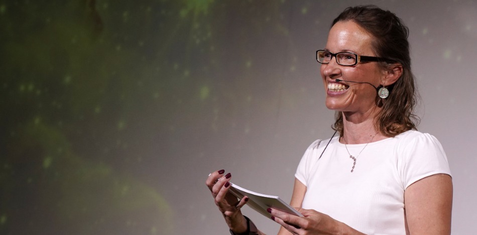 Eine Frau, die Moderationskarten in der Hand hält und in ein Publikum lacht. 