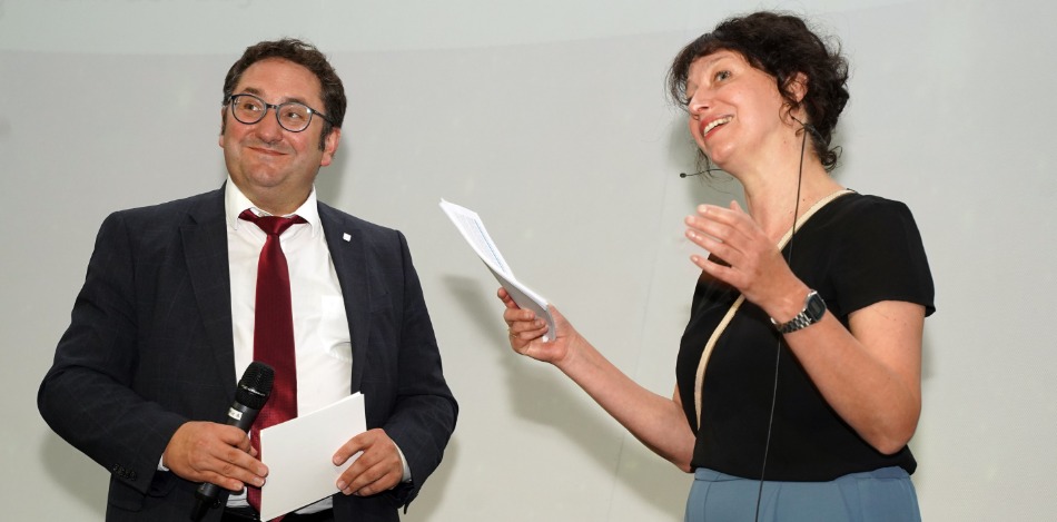 Zwei Personen stehen und interagieren miteinander. Die Person links hält ein Mikrofon und ein Blatt Papier, die Person rechts hält mehrere Blätter und scheint zu sprechen.