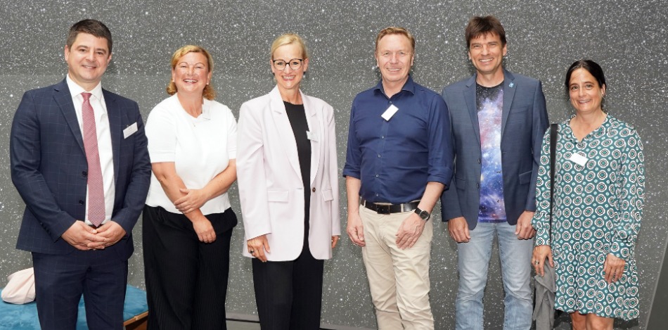 Sechs Erwachsene, drei Männer und drei Frauen, stehen vor einer Wand und lächeln in die Kamera.