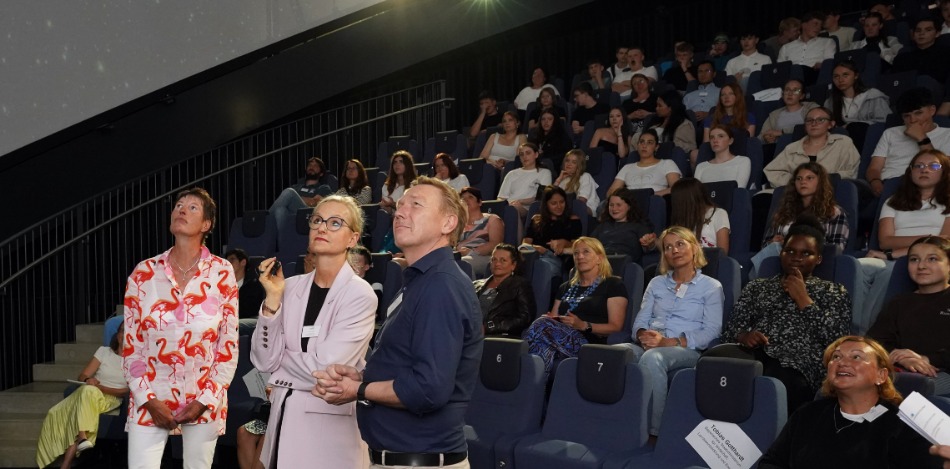 Ein Planetarium ist mit Menschen gefüllt, die in Reihen sitzen und gespannt an die Decke schauen. Drei Personen stehen vorne und sprechen zum Publikum, das aufmerksam zuhört.