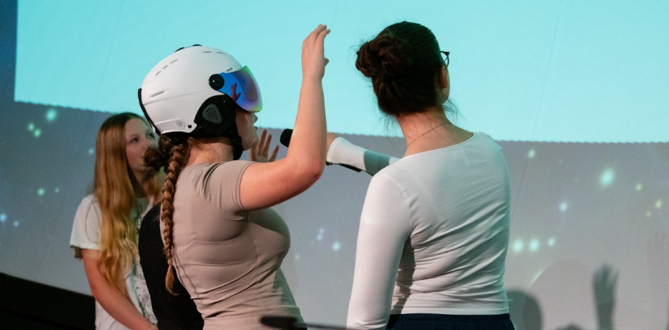 Eine Schülerin mit Helm spricht in ein Mikrofon, während zwei weitere Schülerin aufmerksam zuhören.