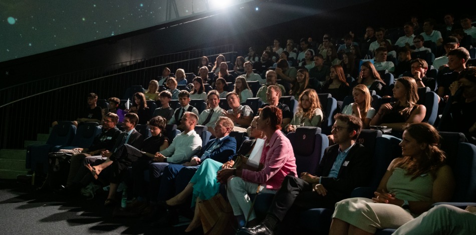 Ein Planetarium ist mit Menschen gefüllt, die in Reihen sitzen und gespannt nach vorne schauen.