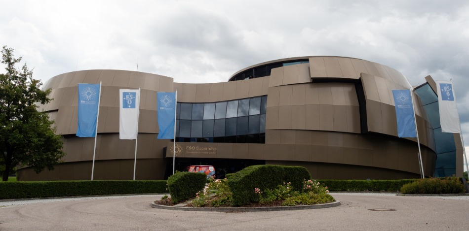 Ein großes graues Gebäude in futuristischem Stil mit wehenden Fahnen davor und Bäumen links und rechts