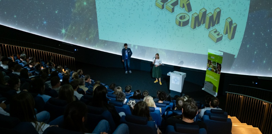 Eine Sprecherin und ein Sprecher stehen mit Mirkos in der Hand vor einer großen Leinwand im vollen Planetarium und begrüßen das Publikum.