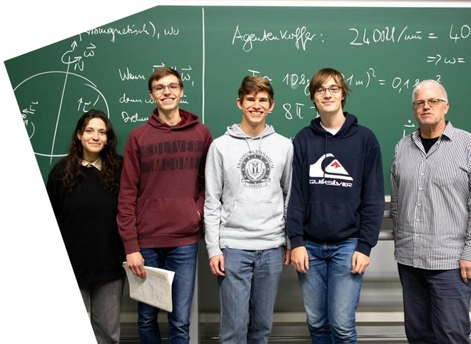 Eine Schülerin steht neben drei Schülern und einem Mann vor einer Tafel, auf der etwas geschrieben steht, und lächeln in die KameraVier Schüler, die Plakate in der Hand halten und mit drei Erwachsenen für ein Foto posieren