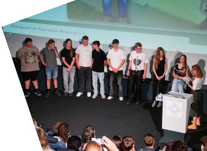 Auf dem Bild steht eine Gruppe junger Menschen auf einer Bühne und präsentiert sich vor einem Publikum.