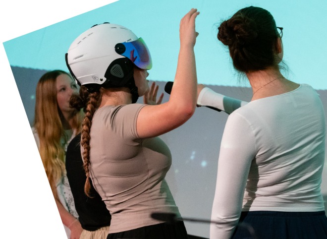 Zwei Personen mit erhobenem rechten Arm stehen vor einer Leinwand, eine davon trägt einen weißen Helm und eine VR-Brille; im Hintergrund sind zwei weitere Personen sichtbar