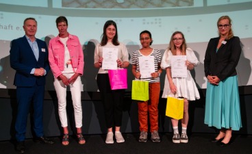 Drei Schülerinnen, die Plakate in der Hand halten und mit drei Erwachsenen für ein Foto posieren